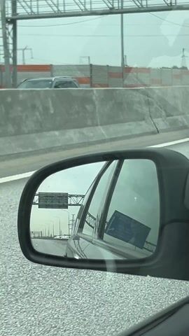 The video shows the side view mirror of a moving car. The car is traveling on a road or highway on a cloudy day. The road is wet. In the mirror, you can see the back of the car and the road behind it. There is a sign visible in the distance. On the side of the road, there is a concrete barrier with orange and white stripes. Power lines are also visible in the distance.