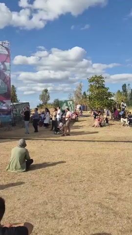 A sunny outdoor gathering is depicted. The ground is covered in dry, yellow grass. Several people are scattered across the scene, some sitting in lounge chairs, others standing in small groups. Two white food trucks are visible in the background, with people gathered around them. A large red and white sign is partially visible on the right side of the frame. The sky is blue with scattered white clouds. The overall impression is of a casual outdoor event or festival.