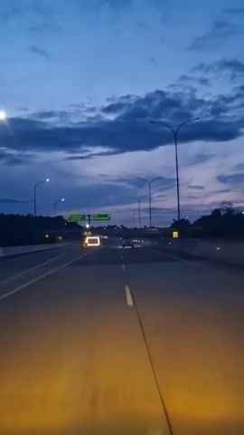 The video shows a first-person perspective of driving on a highway at dusk. The sky is a deep blue with dark clouds. There are streetlights and highway signs visible. The road is asphalt with white lane markings. The headlights of the vehicle illuminate the road ahead with a yellow glow. There are other vehicles on the highway, some appearing as distant lights. The overall scene is one of travel on a highway in low light conditions.