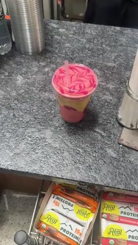 A cup filled with a layered pink and yellow drink sits on a gray countertop. The drink is topped with a pink swirl design on the lid and a straw is inserted. Other items are visible on the counter, including a stainless steel container, a jar, and packaged protein snacks below. Someone wearing a dark shirt is partially visible in the background. The camera focuses on the drink, highlighting its colors and design.