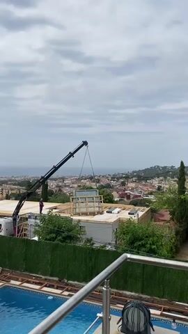 A crane is lifting a large, glass-encased structure, possibly a hot tub or pool, onto the roof of a building under construction. A person in a purple shirt is standing on the roof, observing the operation. The building has a wooden frame. In the foreground, a swimming pool is visible. The background features a town with red-tiled roofs and the ocean under a cloudy sky.
