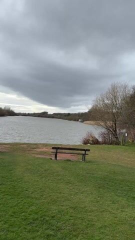 The video shows a lake on a cloudy day. The lake is surrounded by trees and grass. There is a bench and a trash can on the grass near the lake. The wind is blowing, creating ripples on the water. A red buoy stands near the lake. A path runs along the side of the lake.