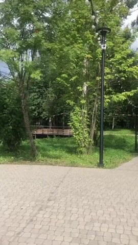The video shows a pathway in a park. The pathway is made of light-colored paving stones. On the left, there is a tall lamp post with multiple lights. There are trees with green foliage on both sides of the pathway. In the distance, there is a small bridge. In the first clip, a few people are seen walking on the pathway towards the bridge. In the second clip, an elderly woman wearing a hat and glasses is sitting on a bench. She is wearing a light pink top and light brown pants. Behind her, there is a playground with swings and a red structure. In the third and fourth clips, the pathway continues with trees and some park features such as arches and benches. The sky is cloudy.