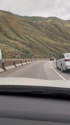 The video is a first-person view from inside a vehicle traveling on a highway. The highway curves to the left, bordered by a concrete barrier and steep, grassy hills on the left. Other vehicles are visible on the road, including a silver car in the foreground on the right lane, followed by a dark-colored sedan and a blue truck in the distance. The sky is overcast.