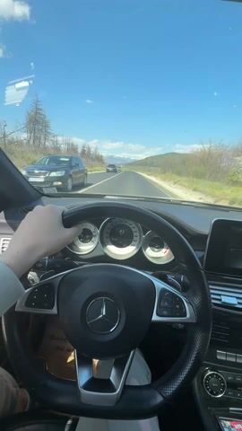 The video depicts the interior of a Mercedes car, focusing on the driver's perspective. The driver's left hand is visible on the steering wheel. The car is moving on a two-lane highway under a clear, blue sky with scattered clouds. The speedometer reads approximately 87 km/h. Distant snow-capped mountains are visible on the horizon. Burned trees line the left side of the road. Other cars are on the road ahead. The dashboard and interior of the car are visible. Music is playing.