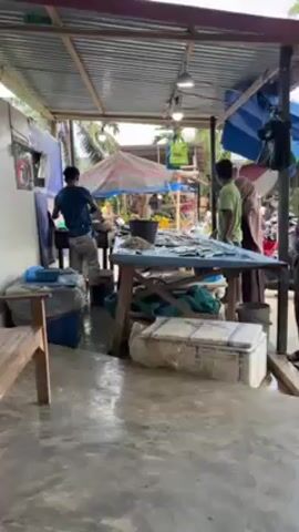Here's a detailed description of the video:

The video captures a lively scene at what appears to be an outdoor fish market. People are gathered around a large table covered with fish, possibly making purchases or examining the selection. A man in a blue shirt is actively working, seemingly preparing or sorting the fish. Several other people are standing around, observing the activity and interacting with each other.

The setting is a covered outdoor area with a corrugated metal roof supported by wooden beams. There are various containers, coolers, and other items scattered around the space. Outside the covered area, there is a street with parked motorbikes and people driving by on scooters. Further in the background, there are houses and greenery.

The camera pans slightly to show different parts of the scene, revealing more details about the market setup and the surrounding environment. There's also a counter with various colorful plastic containers filled with what appear to be food items. Overhead, the roof structure is covered with various packages and hanging items.