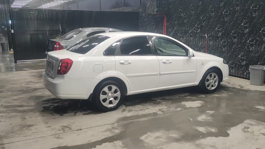 A white, four-door sedan is parked inside a car wash. The car is clean and wet. To the left of the white car, partially visible, is another gray sedan. The floor is wet concrete and water is pooling in places. The background includes a black tarp-like material and a wall with a dark, textured design. A gray bucket is in the right foreground.