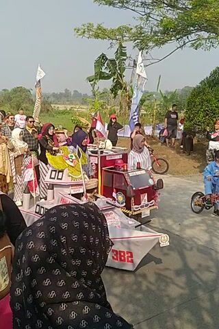 The video captures an outdoor scene with a procession or parade. A group of people, some in traditional attire, are gathered around a decorated vehicle that appears to be a modified cart or small float. The vehicle is adorned with flags, banners, and possibly religious or cultural symbols. There are also individuals on a bicycle with a child dressed in blue. The environment suggests a rural or semi-rural setting with greenery and a pathway or road. The overall impression is one of a festive or celebratory event.
