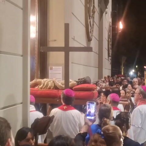 A crowd of people is gathered in front of a building at night. A statue of Jesus Christ lying on a red platform with a wooden cross is being carried out of the building. A group of people in white robes and pink skullcaps walk with the statue. Some people are holding tall lit torches. Many people in the crowd are taking pictures or videos with their phones. The building has stone columns and a window above the entrance. A traffic light is visible in the background.
