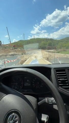 The video shows the interior of a van on a sunny day. The camera is positioned behind the steering wheel, looking out the windshield. The van is on a dirt road that appears to be under construction or leading to a construction site. There is a river or stream next to the road, and a temporary bridge or crossing is visible. Construction equipment, including a crane, is in the background. The dashboard of the van is visible, showing the speedometer and other gauges. A smiley face air freshener hangs from the rearview mirror. The overall environment appears to be rural or undeveloped.