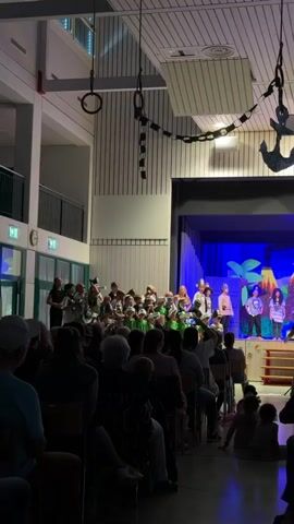 The video shows an indoor performance event, likely a school play, taking place on a stage. The stage is decorated with a blue backdrop featuring palm trees and what appears to be a pirate flag with a skull and crossbones. Several children are on stage, dressed in costumes, some appearing to be singing or acting. In the foreground, an audience of adults and children are seated, watching the performance. The environment appears to be a gymnasium or auditorium, with visible ceiling structures, lighting, and walls. The overall atmosphere suggests a community event or a school performance.