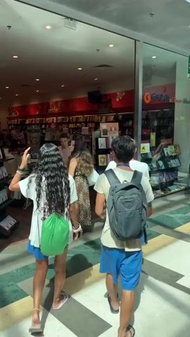 A group of four young people enter a brightly lit bookstore. The two individuals in the foreground, a female with long dark curly hair and a male with a backpack, walk into the store from the right side of the frame. The female is wearing a white t-shirt, blue shorts, and carrying a green bag. The male is wearing a light-colored t-shirt, blue shorts, and a black backpack. Behind them, two other individuals, a male and a female, are visible further inside the store. The male is wearing a light pink t-shirt and the female is wearing a leopard print dress. The bookstore is filled with shelves of books, a large red accent wall with graphics, and display stands. The flooring appears to be a light-colored tile. The individuals are in motion, entering the space.