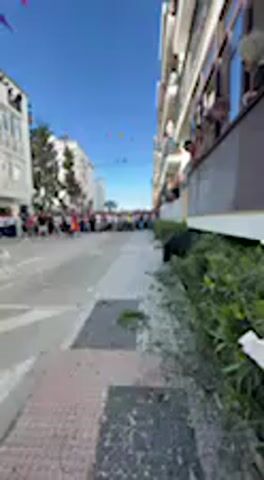 This daytime video captures a vibrant street scene, likely in Vejer de la Frontera, Spain, under a clear blue sky. A large black bull is prominently featured, running at speed along a paved street, moving from the right side of the frame towards the left.

Multi-story white buildings with multiple balconies line both sides of the street. Numerous people are visible on the balconies, leaning over and watching the activity below. Further down the street in the distance, a large crowd of people is gathered, suggesting a public event or celebration.

Colorful decorative flags or bunting are strung across the street overhead, adding to the festive atmosphere. The foreground shows a sidewalk with green foliage along the edge. The bright sunlight indicates clear weather. The audio is dominated by the sounds of a cheering crowd and general outdoor ambience, consistent with a public event. No visible text is present in the video.