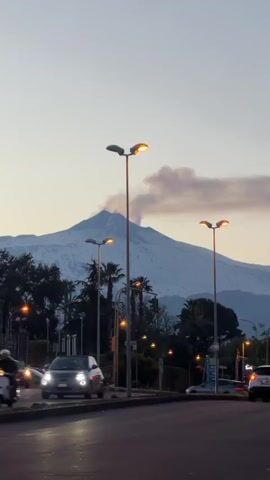 The video captures a scene on a city street with Mount Etna visible in the background. The street is lined with streetlights, trees, and palm trees. Cars are driving on the road. Mount Etna is partially covered with snow, and there is smoke or steam rising from its peak. A gray Alfa Romeo car is prominently featured as it drives away from the camera.
