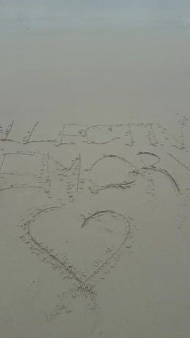 The video shows a wide shot of a sandy beach with the ocean in the background. The text "COLLECTING MEMORIES" is written in the sand, and a heart shape is drawn below it. The waves are gently lapping at the shore. The lighting is overcast, suggesting it may be early morning or a cloudy day.