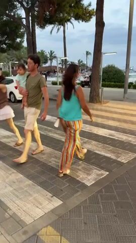 The video shows a person walking across a crosswalk. The person is wearing a turquoise top and striped pants. The crosswalk is made of white stripes on a dark pavement. In the background, there are trees, streetlights, a car, and a body of water with boats. The sky is cloudy. The lighting suggests it is either dawn or dusk.