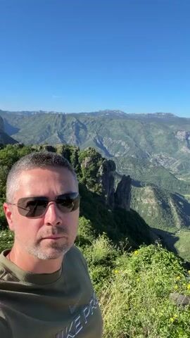 A man wearing sunglasses and a green shirt poses for a selfie. He is positioned in the foreground against a backdrop of lush green mountains and a clear blue sky. The mountainous landscape extends into the distance, with rocky outcrops and valleys visible. The man's face is partially obscured by his sunglasses.
