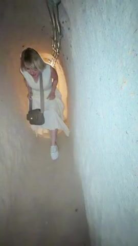 The video captures an exploration of a narrow, low-ceilinged underground tunnel system in Acıgöl, Turkey. The tunnel walls are made of carved rock or earth, illuminated by warm artificial lighting. A person wearing a black top, black pants, and white shoes is seen bending over to navigate the confined passage. This individual points at the textured wall of the tunnel, seemingly highlighting a feature. In the initial blurry frame, another person in a white dress is partially visible, while another individual is seen further down the tunnel in the background. The scene suggests an experience of discovering and exploring an ancient subterranean structure.