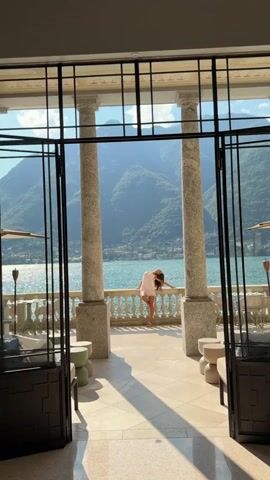 A woman in a light-colored dress stands on a balcony overlooking a lake and mountains. She poses for the camera, then turns and walks away before turning back to face the camera again. The scene is framed by large windows and columns.