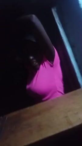 A woman wearing a bright pink shirt looks through a small opening. She has short, dark hair and is smiling. Someone holds a blue phone up to the opening, then moves it. The woman is partially obscured by the opening, which appears to be a window or similar small space. The surrounding area is dark. A wooden surface is visible at the bottom of the frame.