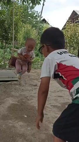 The video shows two young children in an outdoor setting, likely a yard or garden. A baby is seated in a handmade wooden swing, which is suspended from a tree. The baby is smiling and laughing gleefully as they are being pushed on the swing. The baby is wearing a white t-shirt, pink leggings, and light blue socks.

Another child, who appears to be older, is standing in front of the swing and is either pushing it or preparing to push it. This child is wearing a white t-shirt with red and green accents on the sleeves and back, black shorts, and sandals. They are also wearing glasses. Visible text on the older child's shirt includes "ROM" and the number "1" on the back, suggesting it might be a sports jersey.

The background consists of lush green trees and bushes, with a glimpse of a house with a green roof in the distance. The ground is a mixture of dirt and gravel. The lighting suggests it is daytime, likely a sunny day. The overall mood of the video is joyful and playful, with the baby's laughter being a prominent sound. The scene depicts a moment of simple childhood fun.