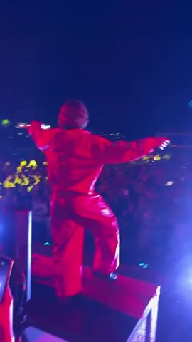 A person in a bright red outfit stands on a raised platform on a stage at night. They are facing away from the camera, towards a large crowd of people. The crowd is illuminated by bright blue and pink stage lights. The person appears to be gesturing with their right arm and holding a microphone to their head with their left. There are speakers and equipment visible on the stage. The sky is dark, indicating it is nighttime.