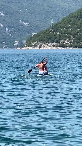 A person is paddleboarding on the water. The person is sitting on the board with their back to the camera, facing a green, mountainous landscape in the distance. The water is blue and choppy. The person is wearing swim trunks. A paddle is visible in their hands.