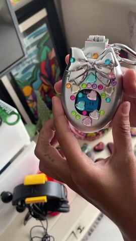 A person's hands are shown holding a compact makeup mirror that is decorated with a silver bow and colorful rhinestones. The mirror is open, revealing a mirrored surface on one side and a black hairbrush on the other. The person is holding the compact in front of a cluttered background that includes artwork, plants, and other objects on shelves. The person's hands have tattoos. The video also shows a close-up of the compact's decorative bow and rhinestones, and then a shot of the hairbrush. The compact is being opened and closed, and the hairbrush is being held up for display.