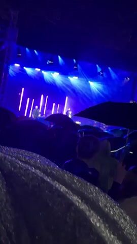 A concert is taking place with bright blue lights illuminating the stage and the audience. The crowd is mostly obscured by umbrellas, suggesting it may be raining or the umbrellas are being used for shade. Several individuals are visible in the foreground and midground, some holding up phones to record or take pictures. The overall atmosphere appears to be energetic and lively, despite the potential for inclement weather.