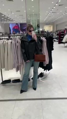 A woman stands in a clothing store taking a selfie in a mirror. She is wearing sunglasses, a blue turtleneck, a black leather jacket, light blue flare jeans, and black high-heeled shoes. A brown bag hangs over her shoulder. The store is well-lit with many racks of clothing in the background. Other customers and store employees are visible in the distance. The floor is light-colored and reflective.