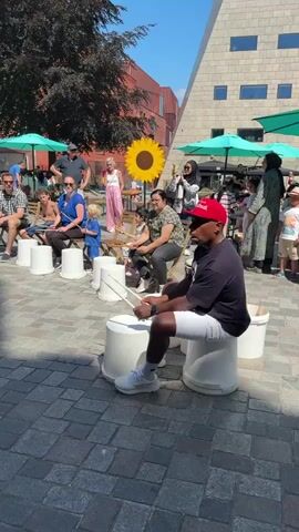 This is a wide shot of an outdoor event, possibly a festival or street performance. A group of people, varying in age, are seated on white buckets in a row, facing towards the viewer. They are all holding drumsticks, and some are actively drumming on their buckets. In the foreground, a young girl in a pink dress is also seated on a bucket, holding drumsticks. In the background, there are more people standing, some sitting at tables under umbrellas, and a large sunflower decoration. The ground is paved with cobblestones, and there are buildings and trees visible in the background. The overall atmosphere appears to be festive and engaging, with the performers interacting with the audience through their drumming.