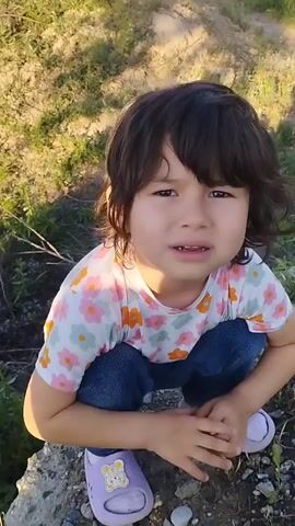 The video opens with a close-up of a young child, likely a girl, squatting on the ground. She has dark, shoulder-length hair with bangs and is wearing a white t-shirt with a colorful floral pattern, blue jeans, and light purple clogs decorated with cartoon bear designs. Her expression is somewhat subdued or thoughtful. The background immediately behind her is blurred green grass and dirt.

The camera then pans down to show the grassy ground before panning up to reveal a wider outdoor scene. In the distance, a single-story, light-colored building with a dark, possibly tiled roof and multiple rectangular windows with white frames is visible. An open area in front of the building suggests a garage or workshop, where a few people are seen moving or working. Another individual is seen walking outside the building to the right.

The foreground of this wider shot is a large, unkempt grassy field with patches of bare dirt and some dry brush. The sky is clear and blue, indicating a sunny day. The overall setting appears to be a rural or semi-rural area in Mingchinar, Uzbekistan. No visible text is present in the video.
