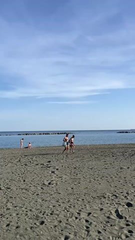 The video depicts a beach scene under a partly cloudy blue sky. The sand is a dark, grainy grey, marked with footprints and the impression of wind-blown patterns. In the foreground, two figures, a male and a female, are walking across the sand. The male is wearing blue patterned swim trunks, while the female is in a two-piece bathing suit. Further along the beach, two more people are visible; one person is standing at the edge of the water, and another is sitting on the sand. The water is a calm, dark blue, marked by a line of rocks stretching into the distance to the left and right, forming breakwaters. A large ship is visible on the horizon. The video is taken from a static position at ground level.