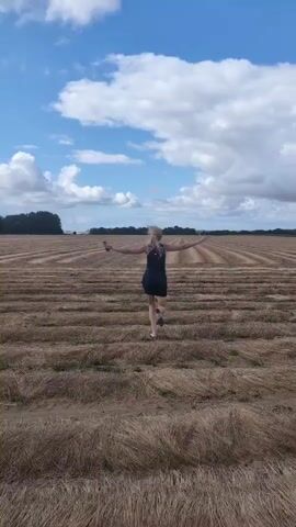 A woman with blonde hair wearing a dark dress and white shoes is running away from the camera through a harvested field. The field is filled with rows of dry, brown stubble. The sky above is blue with scattered white clouds. The woman has her arms outstretched to her sides. The camera is filming from behind her at a slight downward angle.