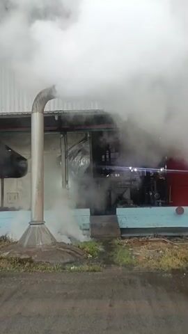 A vertical video shows a large industrial facility with a tall, silver smokestack emitting a thick plume of white smoke. The smokestack is curved at the top. To the right of the smokestack, a building's facade is visible, painted in sections of white, light blue, and red. The lower portion of the building's wall is light blue, with some dirt and grass growing along its base. The ground in front of the building appears to be a dark, paved surface. The smoke billows outwards and upwards, obscuring parts of the building and the machinery visible behind it. The overall scene suggests industrial activity, possibly the release of steam or emissions.