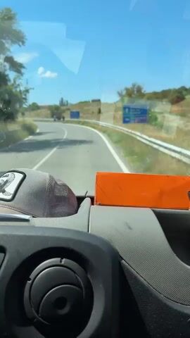 The video shows a view from inside a vehicle, looking out through the windshield. The scene is a sunny day, with a clear blue sky and some light clouds. A paved road curves to the left, bordered by a white line and a metal guardrail. On the right side of the road, there is a blue directional sign with white text. The text is in Bulgarian and reads: "Стремци" (Stremtsi) and "Кърджали" (Kardzhali) with arrows indicating directions. There is also some smaller text that is difficult to read.

On the left side of the road, a group of people are standing by the roadside. There are at least four individuals, some appearing to be looking at something or someone off-camera. The perspective suggests the vehicle is moving, indicated by the slight blur of the background and the motion blur on the people.

Inside the vehicle, the dashboard is visible in the foreground. A gray baseball cap with a black emblem of a bird is resting on the dashboard. An orange rectangular object is also visible. The overall impression is of a casual journey on a warm, bright day in a rural or semi-rural setting. The activity appears to be a road trip, possibly with a stop or a point of interest being observed by the people on the roadside. The presence of the directional sign suggests a specific geographical location within Bulgaria.