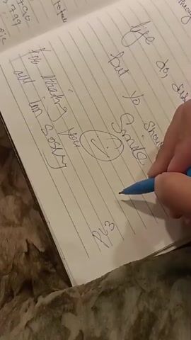 A hand is writing on a lined notebook with a blue pen. The notebook is open to a page with blue lines and some writing in cursive blue ink. Some of the visible text reads "life do, die", "but yo Smile Should", "to Thinking You", "I'm Sorry", and "PLZ". The background appears to be a textured surface, possibly a table.