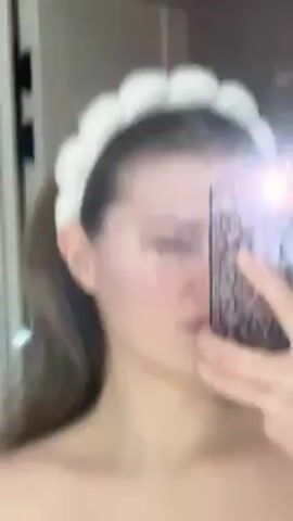 A woman with light skin and long brown hair pulled back into a ponytail poses for a selfie in front of a mirror. She is wearing a puffy white headband. Her expression changes slightly as she looks at the camera. She holds a phone with a floral-patterned case up to the mirror, causing glare. The background shows a white door and tile.
