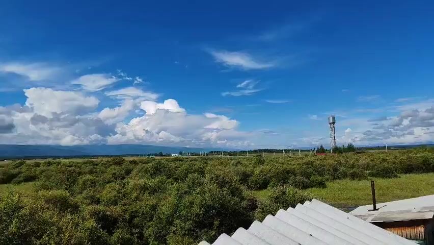The video captures an aerial view of a rural landscape under a bright blue sky with scattered white clouds. In the foreground, the corrugated metal roofs of several rustic wooden structures are visible, including what appears to be a small house with a porch and a smaller shed-like building. A wooden fence encloses parts of the property. Beyond the immediate structures, a vast expanse of green grassland stretches out, dotted with bushes and trees. In the mid-ground, a tall, slender water tower stands prominently against the horizon. Further in the distance, a cluster of houses with various roof types indicates a small village or settlement. The overall impression is one of a tranquil, open countryside scene.