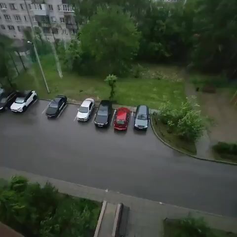 The video shows a residential area with apartment buildings, trees, and a parking lot. There are several cars parked in the lot, including a white car, a black car, a silver car, a black car, a red car, and a gray car. The ground is wet, indicating it may be raining or has recently rained. The sky is overcast. There is a playground visible in the background.