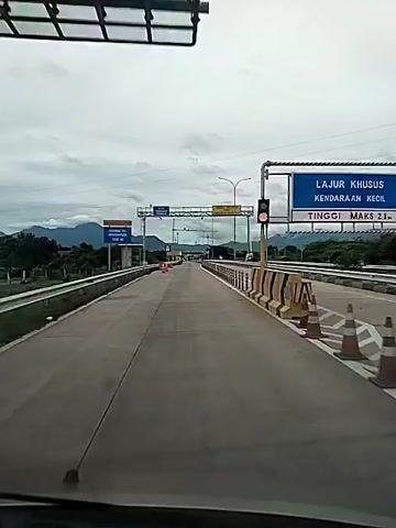 The video depicts a view from inside a vehicle, likely a car, traveling on a highway or toll road. The road is straight and paved with gray concrete. It is divided into lanes. There are guardrails along both sides of the road, and barriers made of black and white posts in the middle of the road. Overhead signs provide information, including one that indicates an automatic weighing area.  Traffic appears light.  The sky is overcast with a thick layer of gray clouds. In the distance, mountains are visible.  The camera remains mostly still, capturing the forward motion of the vehicle.