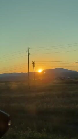This is a vertical video shot from inside a vehicle, looking out at a landscape during sunset. The sun is low on the horizon, partially obscured by a mountain range in the distance, casting a warm orange glow. The sky transitions from a pale orange near the sun to a soft teal at the top. In the foreground, there's a field of tall grass, some of which appears slightly hazy, possibly due to dust or mist. Two utility poles with power lines are visible, one closer to the foreground and another further back. The lower left corner of the frame shows a small portion of the vehicle's side mirror, reflecting some of the light. The overall impression is of a serene, rural setting at dusk.