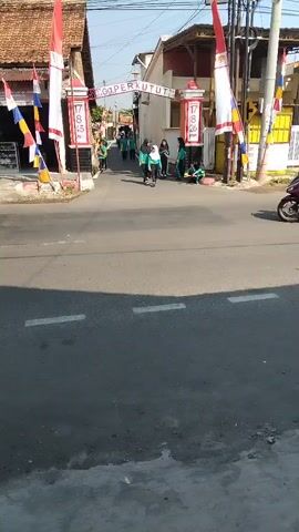 The video shows an outdoor street scene with several people and a motorcycle. A banner hangs across the street with text that appears to be "Go PERKUTUT" and the numbers "17 8 45" and "17 8 46". Several people, mostly young, are gathered under the banner. One person is riding a motorcycle, carrying another person on the back, and passes under the banner. The motorcycle and passengers are moving from right to left across the frame. Red and white flags are flying from poles on either side of the street, and there are other decorations. The scene appears to be a residential or small commercial area. The weather is sunny.