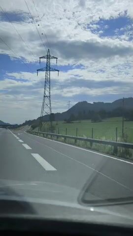 A dashboard camera view shows a car driving on a highway. On the right side of the road, there is a large metal electricity pylon. Beyond the pylon, there is a grassy field bordered by a fence and then a line of trees on a hill. The sky is blue with some white clouds. There are road signs along the highway. The car is driving on a paved road with white lines marking the lanes.