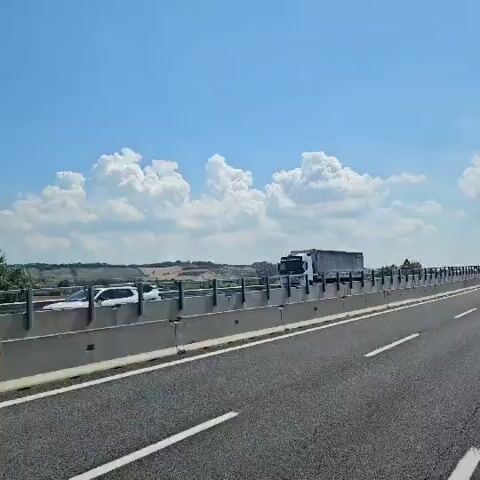 The video shows a view from inside a vehicle traveling on a highway. The sky is blue with scattered white clouds. On the left side of the road, there is a barrier, and beyond that, a truck is visible. The road has multiple lanes marked with white lines. The vehicle continues to move forward, with the scenery of fields and trees passing by on the right side of the road. Power lines and some structures are visible in the distance. The camera angle is stable, suggesting it is mounted.