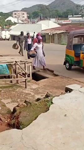 In Gwong, Nigeria, a group of people are gathered near a street with a broken pavement. A woman in a white and purple patterned dress with a headscarf is stepping into a brightly colored auto-rickshaw, possibly a tuk-tuk, which is parked on the right side of the frame. She is holding a large black bag. Behind her, another woman in a red top and black headscarf, also holding a black bag, appears to be helping her. Two men are walking in the background, one carrying a woven basket. A younger woman in a pink top and dark pants is also present, standing near the men. The scene appears to be a marketplace or a busy street with various buildings and a white van visible. The weather seems overcast, and the landscape suggests a hilly terrain in the distance. The broken pavement and exposed drain in the foreground highlight a rough infrastructure. The overall atmosphere is that of everyday life and movement in a Nigerian town.