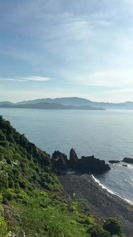 The video shows a coastal landscape. In the foreground, there is lush green foliage and a rocky shore. The sea extends to the horizon, with several islands visible in the distance. A few small boats are scattered across the water. The sun is shining brightly, casting a shimmering reflection on the surface of the sea. The sky is clear with a few scattered clouds.