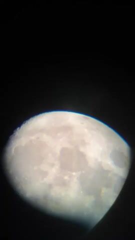 The video shows a close-up, slightly blurry view of the moon. The moon is illuminated, revealing craters and surface features. The background is black. The image is framed by a dark vignette, suggesting it was taken through a telescope or camera lens. There are no people or animals visible. The lighting suggests it is nighttime.