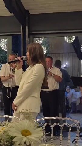A woman in a white suit sings into a microphone. She has long, brown hair and is wearing a white top under her jacket. She is standing in front of a table with white flowers and has her left hand on the microphone and her right hand extended. Behind her, two men play instruments; one plays a clarinet-like instrument and the other plays a trumpet. There are other people in the background, and the setting appears to be an indoor event with a ceiling made of wooden beams and lights.