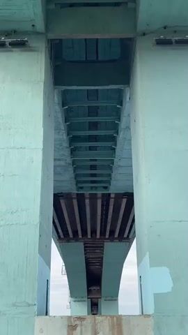 This low-angle shot captures the underside of a large concrete bridge, viewed from directly below and between two massive, light greenish-blue (teal) pillars. Looking up, the intricate support structure of the bridge deck is visible, composed of numerous horizontal and diagonal beams and girders, also painted in similar shades of green and blue, receding into the distance. The overall impression is one of robust, geometric infrastructure. In the background, through the arch of the bridge, a lighter sky is visible, suggesting an outdoor setting during daytime. The lower parts of the concrete pillars show some wear and discoloration. There is no visible activity or text in the image. The scene depicts a large bridge structure, consistent with a location such as Grigor'evskoe, Russia.